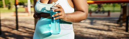 Mujer afroamericana curvilínea en ropa deportiva sosteniendo una botella, rodeada de naturaleza, encarnando la positividad corporal mientras hace ejercicio en un parque.