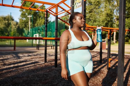Photo for An African American woman in sportswear stands in a park, holding a water bottle, taking a refreshing break from her outdoor workout. - Royalty Free Image