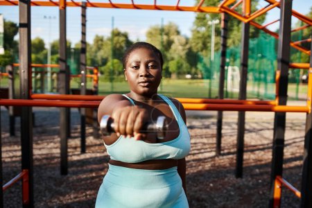 A curvy African American woman in a blue sportswear confidently holds a metal dumbbell in her hands, exuding grace and strength.