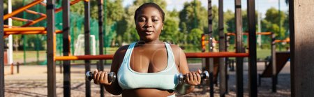 An African American woman with a curvy body is working out in a blue sports bra, holding dumbbells in front of a playground.