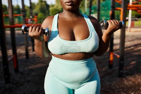 A body-positive African American woman in a blue sports suit confidently lifts two dumbbells outdoors.
