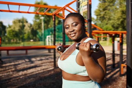 An African American woman in sportswear confidently exercises with a dumbbell in a lush park, embracing body positivity.