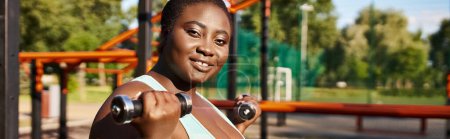 An African American woman in sportswear exercises with dumbbells in a lush park setting.