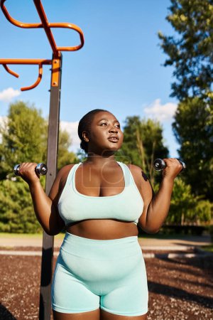 A curvy African American woman in blue workout attire confidently holds a dumbbells in an outdoor setting.