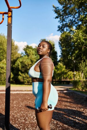 A curvy African American woman in sportswear stands next to a pole in a park.