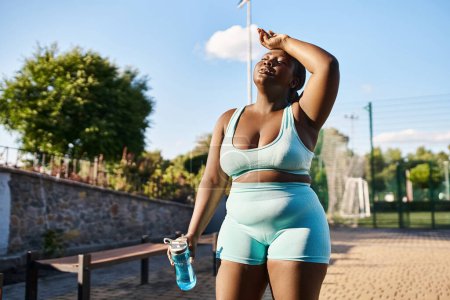 African American woman in sports bra and shorts holding a water bottle, staying hydrated during her outdoor workout.
