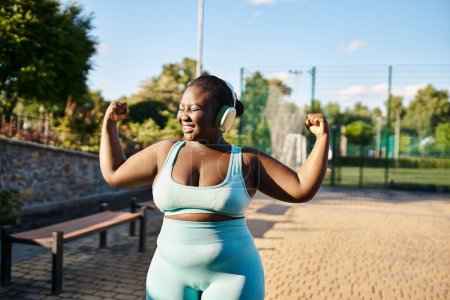 An African American woman in a sports bra and leggings flexes her muscles confidently outdoors.