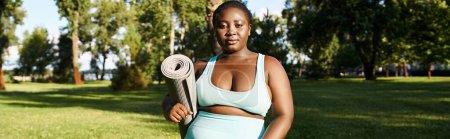 A curvy African American woman in sportswear gracefully holds a rolled up yoga mat in a serene park setting.