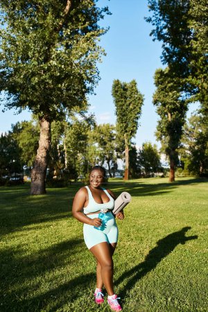 Eine kurvige Afroamerikanerin in Sportbekleidung steht selbstbewusst im saftigen Gras und hält eine Sportmatte in der Hand.