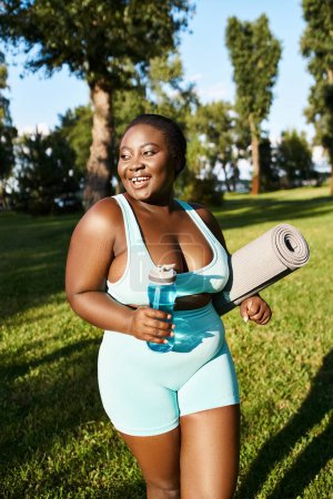 A joyful African American woman in sportswear, curvy and body positive, holds a yoga mat and a bottle of water outdoors.