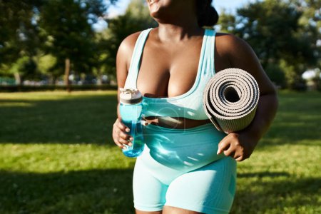 African American woman in blue swimsuit holds yoga mat and water bottle in outdoor workout setting.