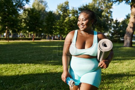 Curvy African American woman in blue sports bra and shorts holding a yoga mat outdoors.