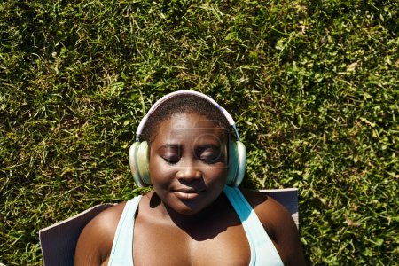 Eine Afroamerikanerin in Sportkleidung entspannt sich in der Natur und hört Musik über Kopfhörer, während sie im Gras liegt.