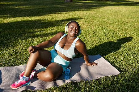 Una mujer afroamericana con curvas en ropa deportiva sentada con gracia en una esterilla de yoga practicando mindfulness al aire libre.