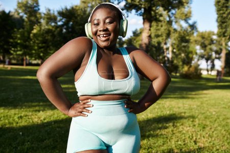 An African American woman in sportswear stands in the grass, embracing body positivity, while listening to music through headphones.
