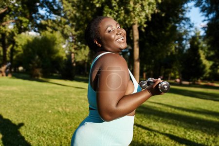 A body-positive African American woman in sportswear holds two dumbbells while exercising in a serene park setting.