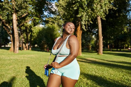 An African American woman in sportswear stands gracefully in a field, surrounded by tall trees, radiating a body-positive aura.
