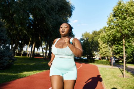 Photo for An African American woman in blue shorts is running with determination on a scenic path surrounded by lush greenery. - Royalty Free Image