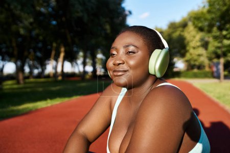 An African American woman in sportswear sits, wearing headphones, immersed in music, embodying body positivity while outdoors.