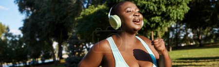 Eine Afroamerikanerin in Sportkleidung mit positiver Körperhaltung steht in einem Park und trägt Kopfhörer.