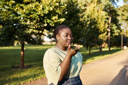 Femme afro-américaine de grande taille se tient sur le bord de la route, bavardant au téléphone dans la chaleur de l'été, embrassant la positivité du corps.