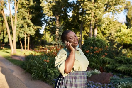 A plus-size African American woman in casual attire enjoying a phone conversation while surrounded by nature in a sunny park.