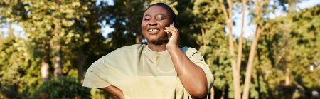 Photo for A plus-size African American woman in casual attire talking on a cell phone while enjoying a sunny day in the park. - Royalty Free Image