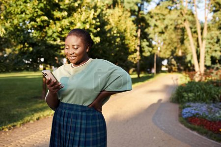 Plus Size Afroamerikanerin in Freizeitkleidung steht in einem Park und schaut an einem sonnigen Tag auf ihr Handy.