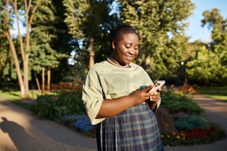 Una mujer afroamericana de talla grande con atuendo casual parada en un parque, absorta en su teléfono celular en un día soleado.