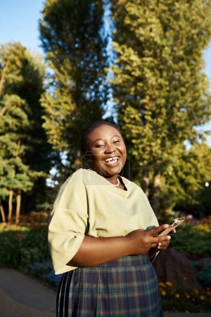 Eine Afroamerikanerin in Freizeitkleidung genießt einen Sommertag im Park und hält ein Handy in der Hand.