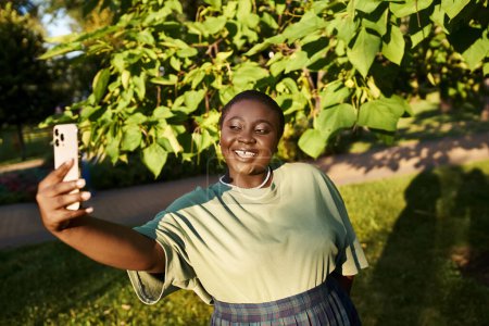 Una mujer afroamericana de talla grande vestida con atuendo casual tomando alegremente una selfie con su teléfono celular al aire libre en el verano.