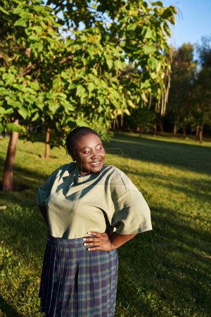 Une femme afro-américaine de taille plus, debout avec confiance dans l'herbe, les mains sur les hanches dans une position corporelle positive.