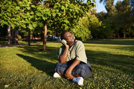Eine Afroamerikanerin in Plus-Size-Größe sitzt im üppigen Gras und ist in der warmen Sommersonne in Musik vertieft.