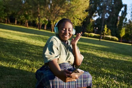 Eine afroamerikanische Plus-Size-Frau genießt Musik, während sie in lässiger Kleidung auf dem Gras sitzt und Körperpositivität umarmt.