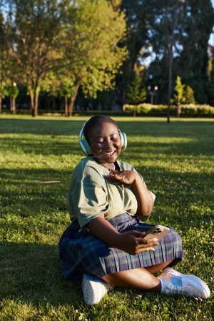 Una mujer afroamericana de talla grande se sienta en la hierba, con auriculares, disfrutando de la música en un día soleado de verano.