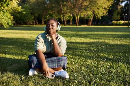 Relaxed woman enjoying music while seated in grassy environment on a sunny day.