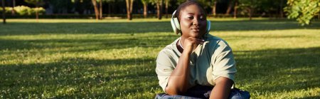 woman in casual attire sits on grass, listening to headphones, surrounded by nature in a peaceful summer moment.