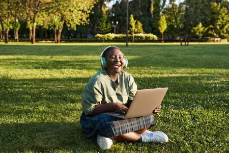 A plus-size African American woman sits on the grass with a laptop, enjoying the outdoors in summer.