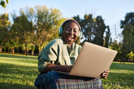 Plus size African American woman sitting in grass with laptop, enjoying outdoor work in summer setting.