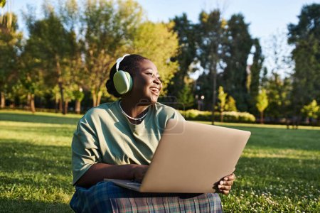 Plus-size African American woman sits in grass, working on laptop in summer. Embracing body positivity.