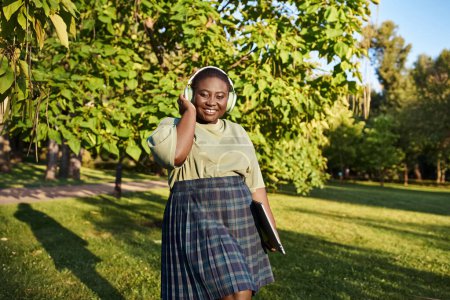 Plus-size woman with African American heritage, dressed casually in a skirt, speaks on a headphones outdoors on a sunny day.