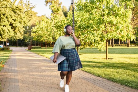 A plus size African American woman in casual attire walks down a sidewalk, on a summer day.