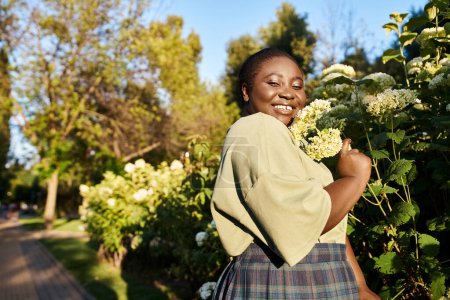 Plus Size Afroamerikanerin steht anmutig neben einem Strauch, der an einem sonnigen Sommertag mit schönen weißen Blumen geschmückt ist.