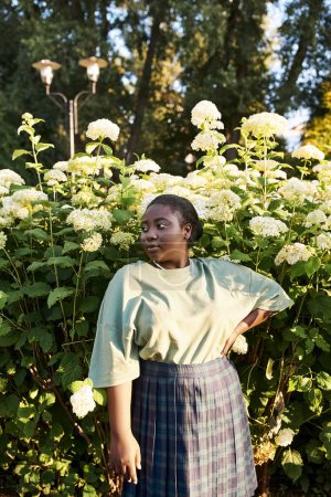 A plus-size African American woman stands confidently in front of a bush with white flowers, embracing the beauty of nature around her.