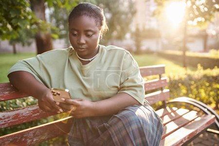 Téléchargez les photos : Une femme afro-américaine de grande taille en tenue décontractée assise sur un banc en bois dans un parc lors d'une journée d'été ensoleillée, embrassant la positivité du corps. - en image libre de droit