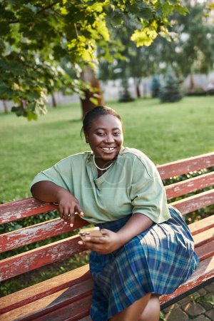 Relaxing plus size African American woman sitting on a wooden bench in a peaceful park setting on a sunny day.