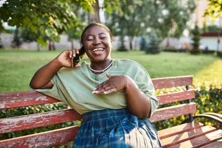 Plus size African American woman in casual attire, sitting on a bench outdoors in the summer, talking on a cell phone.