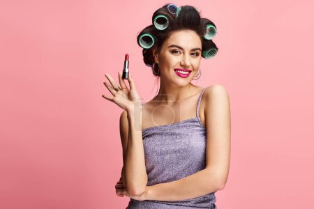 Stylish woman with curlers in hair holding lipstick in front of vibrant backdrop.