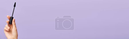 Téléchargez les photos : A womans hand delicately holds a black mascara against a captivating purple backdrop. - en image libre de droit