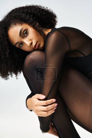 Photo for A young African American woman in black stockings strikes a glamorous pose against a grey studio background. - Royalty Free Image